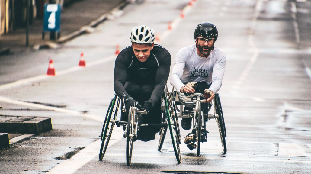 Two men compete in a wheelchair race on a wet urban street, showcasing endurance and teamwork.